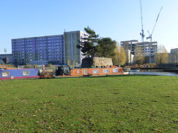 Grassy field with canal boats, trees, and urban skyline under blue sky. — New Islington Marina, Manchester