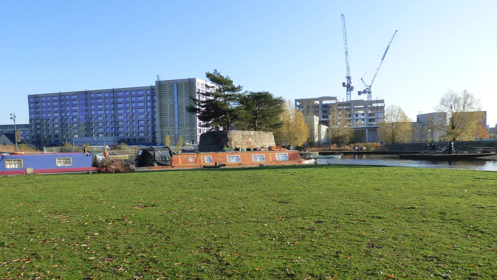 Grassy field with canal boats, trees, and urban skyline under blue sky. — New Islington Marina, Manchester