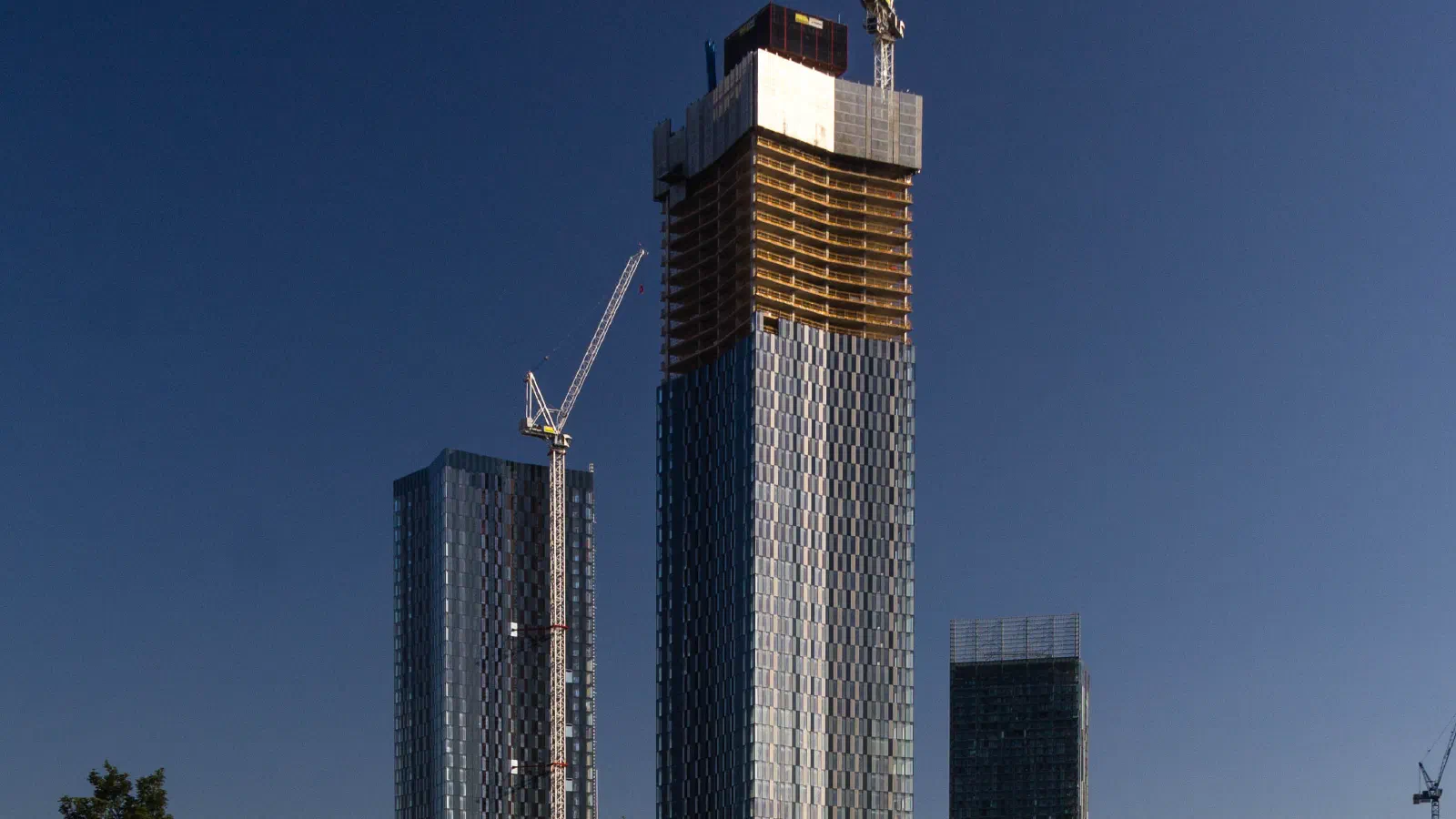 Two craned skyscrapers rise over grassy park against clear blue sky at midday sun. — New Jackson (Deansgate Square), Manchester