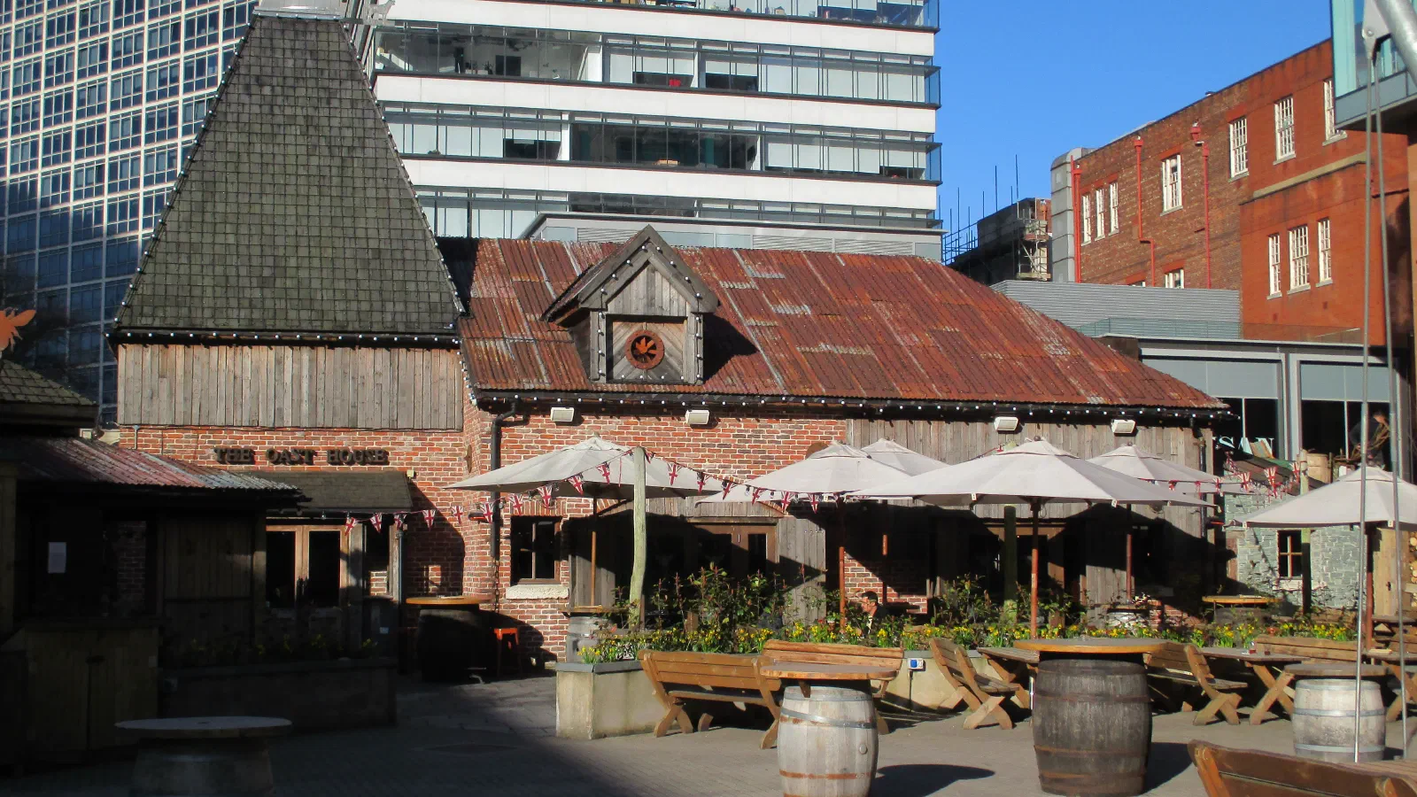 Rustic wooden building with outdoor seating and umbrellas under blue sky. — The Oast House & Courtyard, Manchester