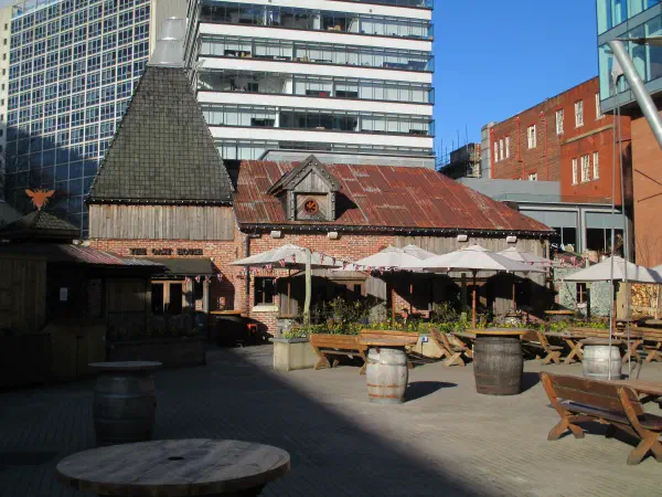 Rustic wooden building with outdoor seating and umbrellas under blue sky. — The Oast House & Courtyard, Manchester