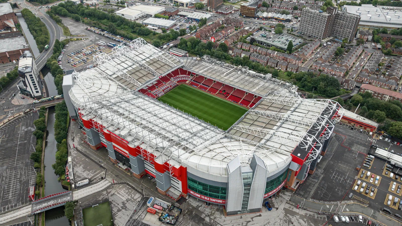 Overhead view of a large football stadium with green pitch, red seating and modern white roof against city backdrop. — Old Trafford Stadium, Manchester