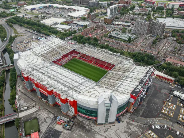 Overhead view of a large football stadium with green pitch, red seating and modern white roof against city backdrop. — Old Trafford Stadium, Manchester