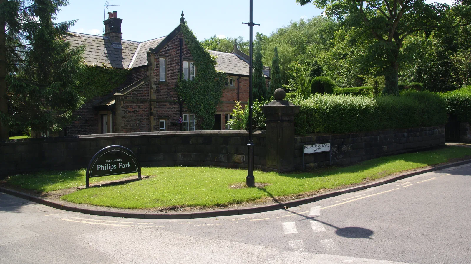 Sign for Philips Park by brick building and trees on sunny day. — Philips Park, Manchester