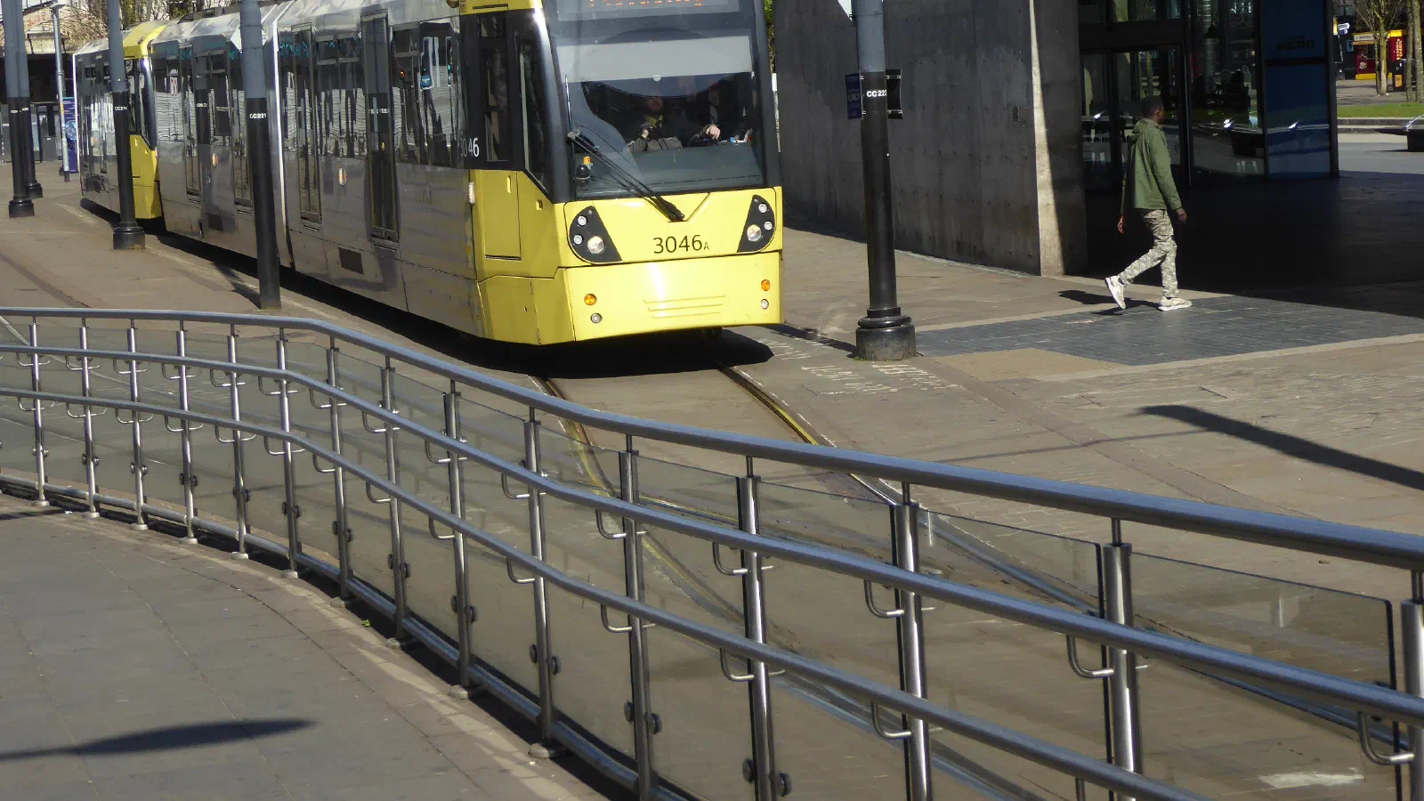 A yellow tram marked 3046x travels along tracks near a building on a sunny day, with trees and pedestrians nearby. — Piccadilly Gardens, Manchester
