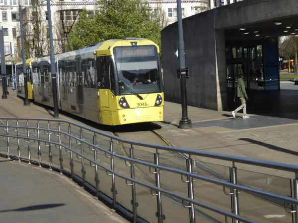 A yellow tram marked 3046x travels along tracks near a building on a sunny day, with trees and pedestrians nearby. — Piccadilly Gardens, Manchester