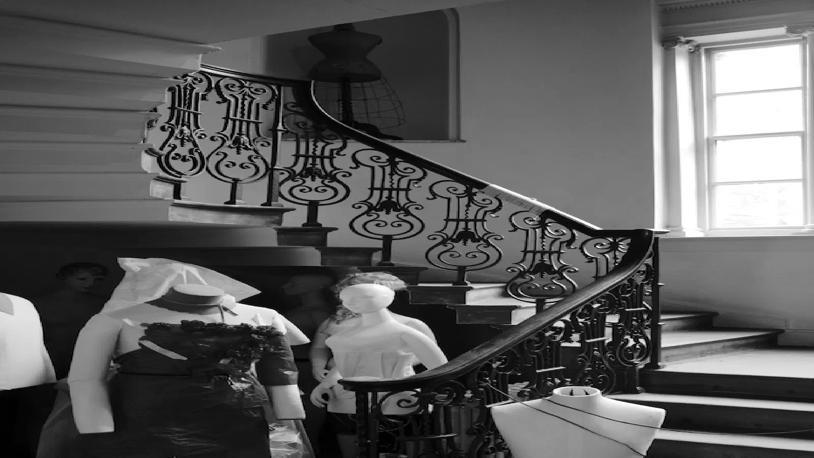 Black-and-white photo shows mannequins on an ornate staircase bathed in natural light from a large window. — Platt Fields Park, Manchester