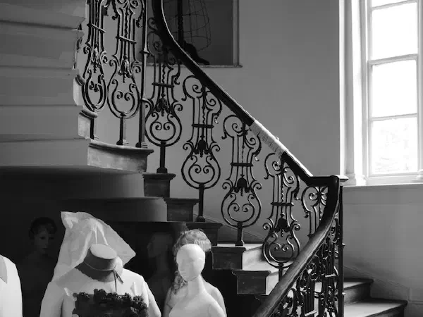 Black-and-white photo shows mannequins on an ornate staircase bathed in natural light from a large window. — Platt Fields Park, Manchester