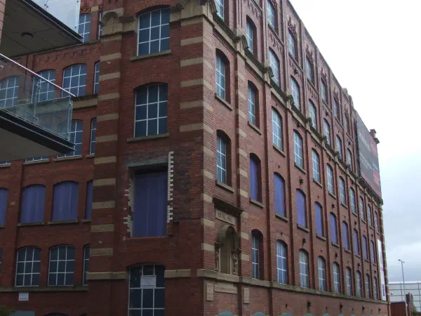 Red brick building on corner, parked blue car beside it, gray sky, glass railings to left. — Royal Mill, Manchester