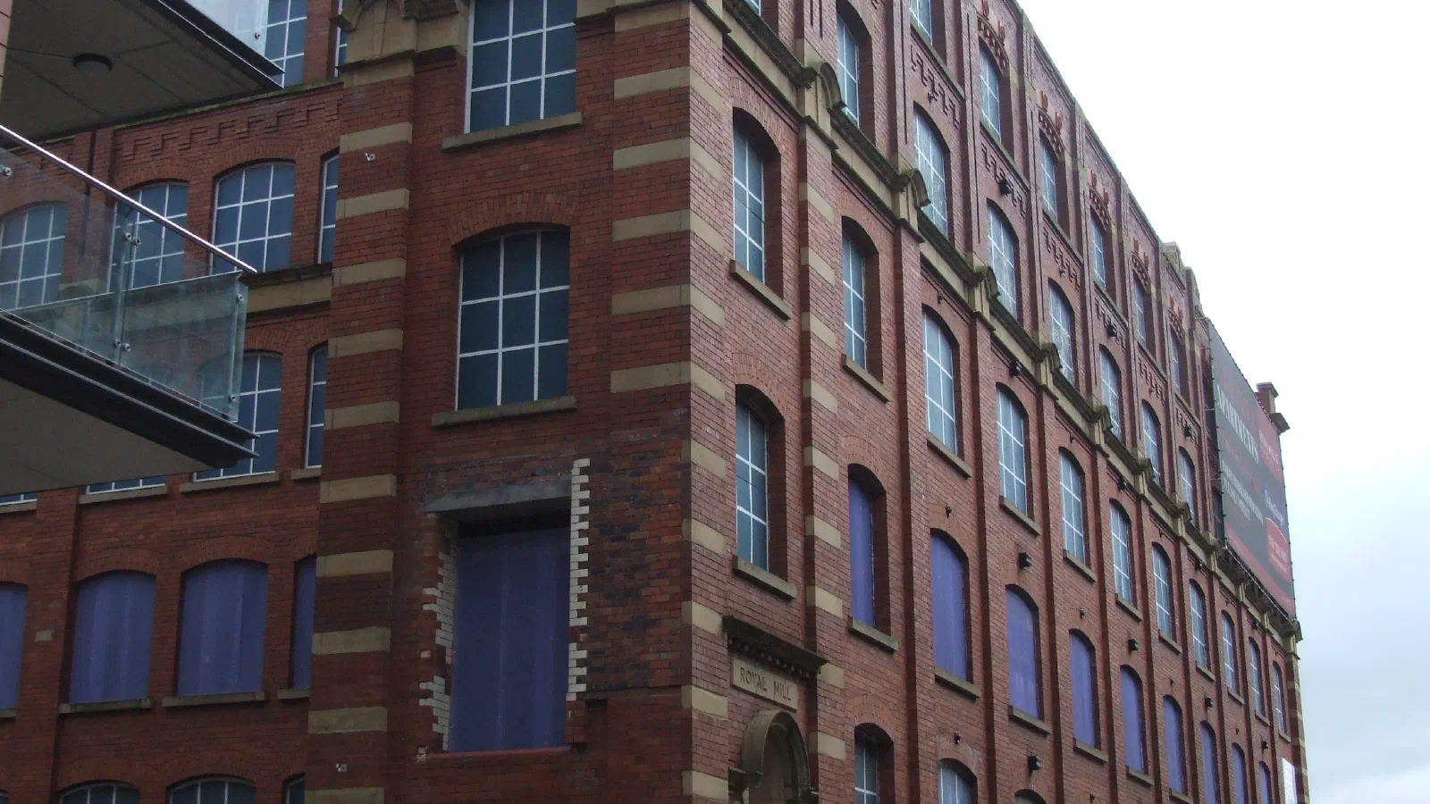 Red brick building on corner, parked blue car beside it, gray sky, glass railings to left. — Royal Mill, Manchester