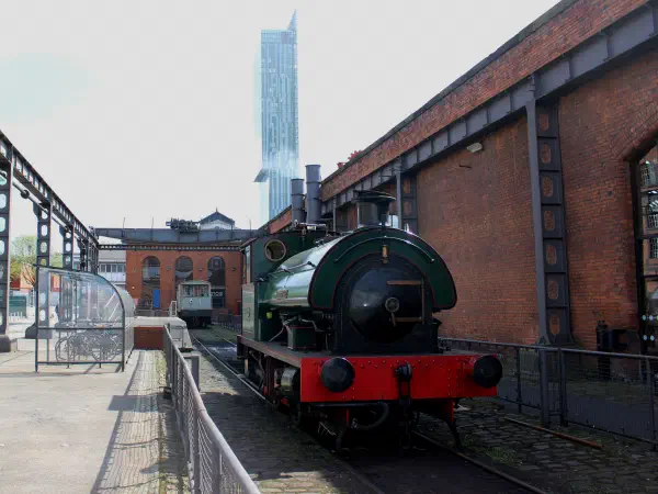 Green steam engine beside red brick building under clear sky near modern skyscraper. — Science and Industry Museum, Manchester