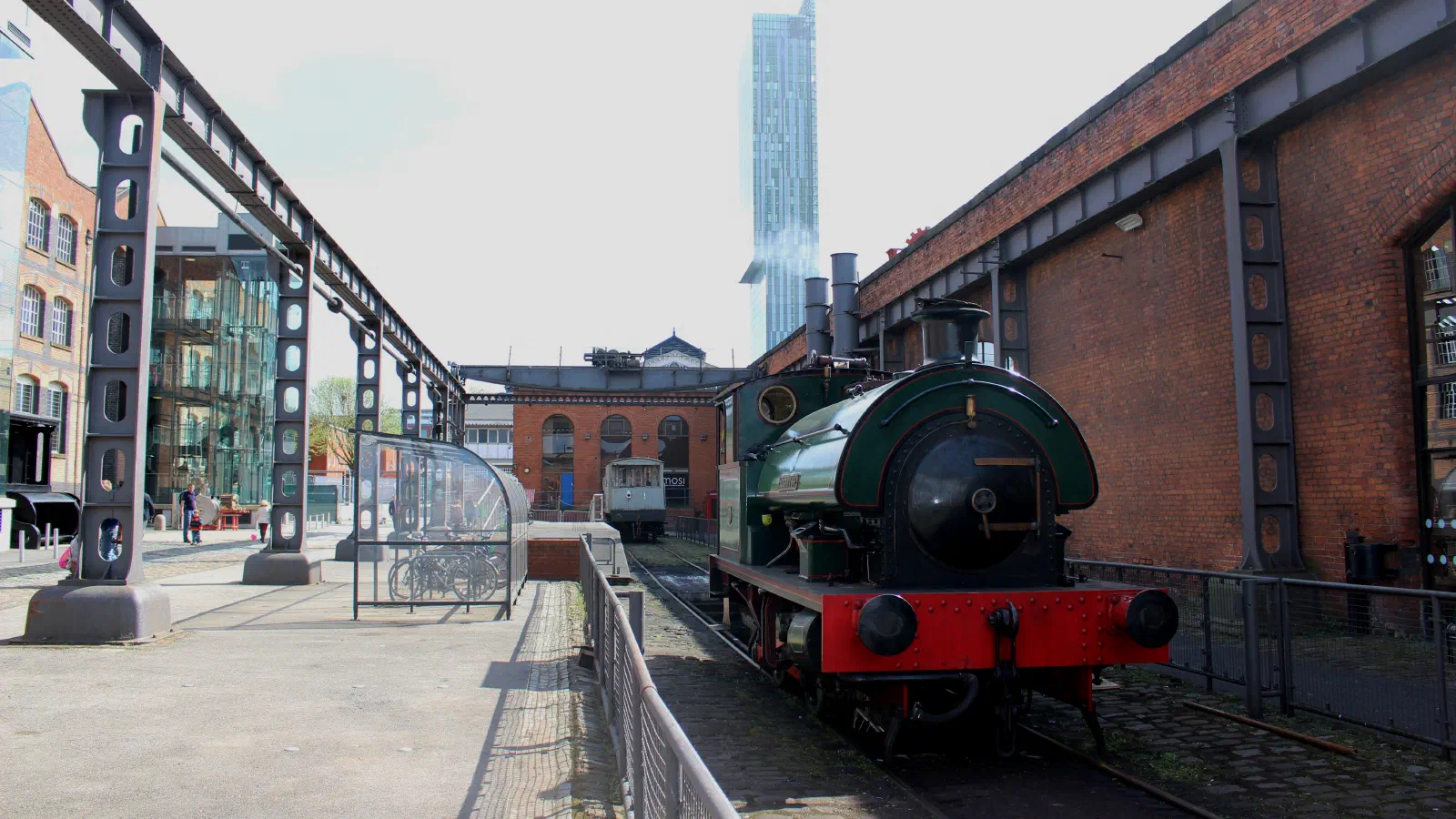 Green steam engine beside red brick building under clear sky near modern skyscraper. — Science and Industry Museum, Manchester
