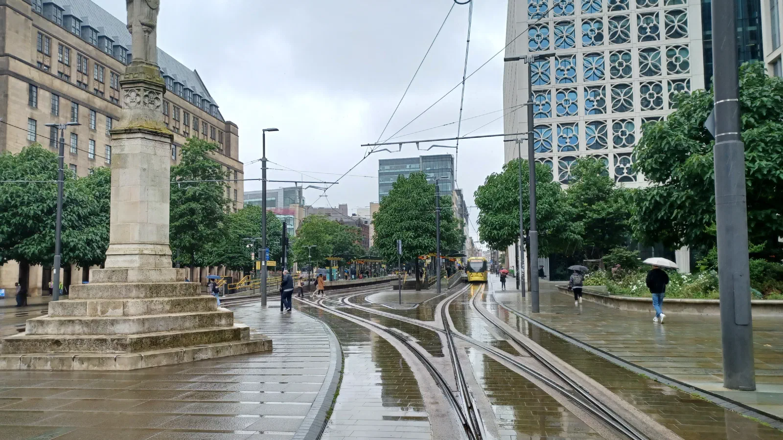 Rainy day on tram tracks; wet pavement reflects light, surrounded by trees and tall buildings. — St Peter’s Square, Manchester