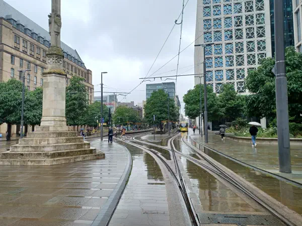 Rainy day on tram tracks; wet pavement reflects light, surrounded by trees and tall buildings. — St Peter’s Square, Manchester