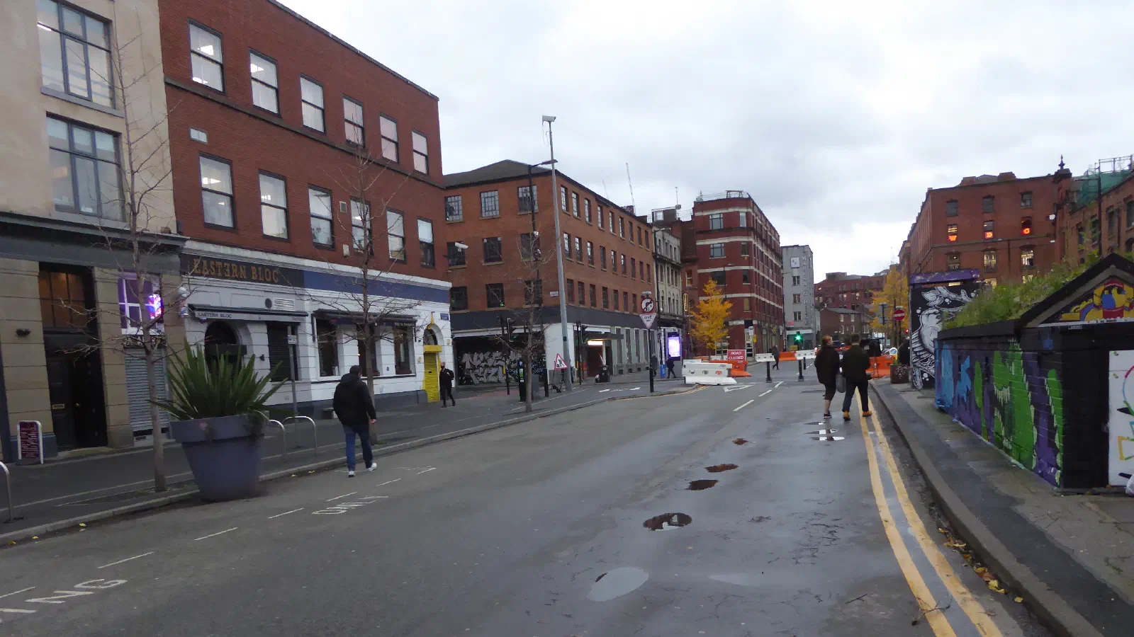 A city street scene with pedestrians, shops like Eastern Bloc, and graffiti-covered walls under an overcast sky. — Stevenson Square, Manchester