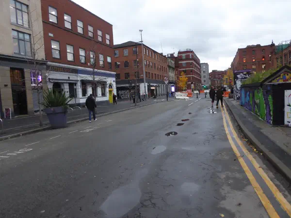 A city street scene with pedestrians, shops like Eastern Bloc, and graffiti-covered walls under an overcast sky. — Stevenson Square, Manchester