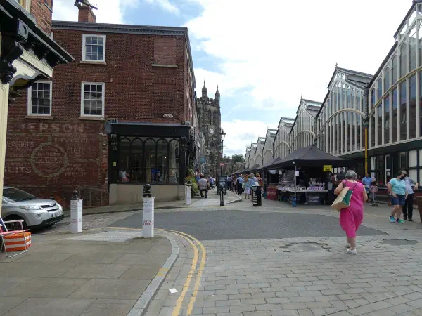 Market stalls under glass roofs line a cobblestone street where pedestrians walk past brick buildings and a silver car. — Stockport Market Place, Manchester