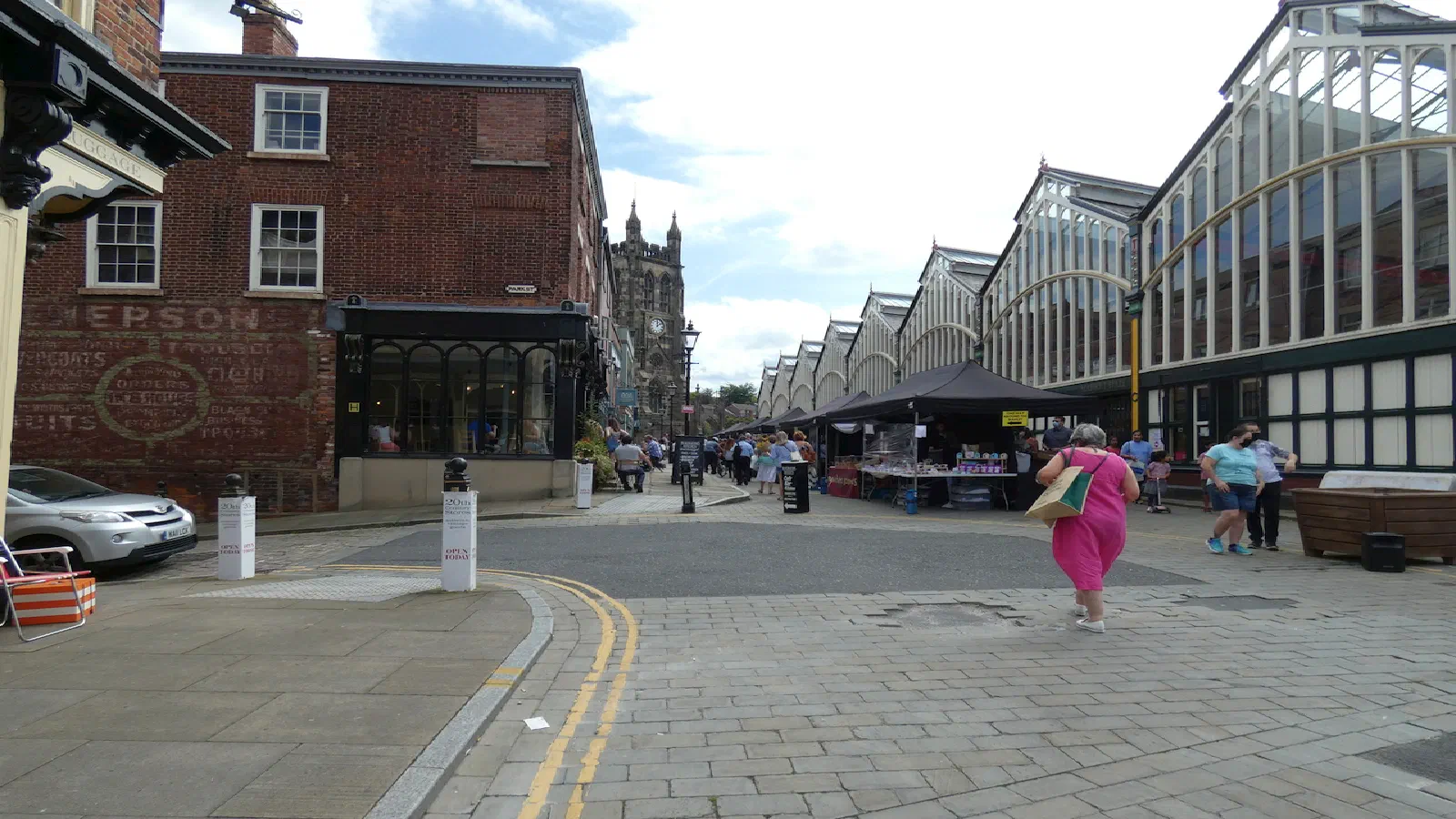 Market stalls under glass roofs line a cobblestone street where pedestrians walk past brick buildings and a silver car. — Stockport Market Place, Manchester
