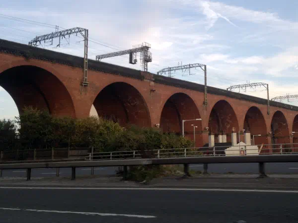 Tall red brick viaduct spans road under clear sky with overhead lines. — Stockport Viaduct, Manchester