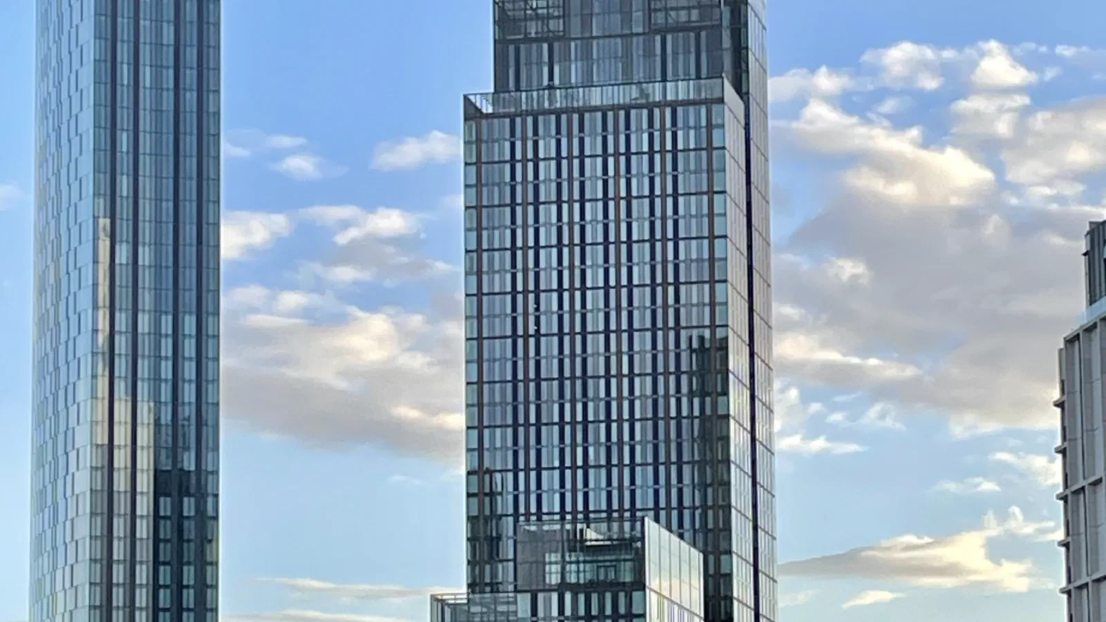 Modern glass skyscrapers rise against a blue sky with scattered clouds on an urban landscape. — The Blade & Three60, Manchester