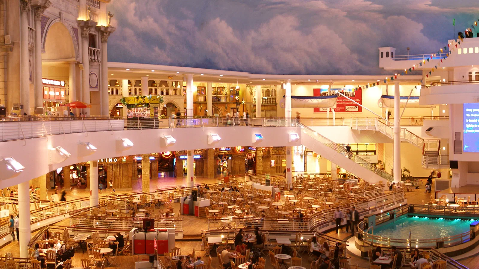 Wide-angle panorama shows a multi-level shopping mall's indoor atrium with restaurants and shops under a painted sky ceiling. — The Trafford Centre (The Orient), Manchester