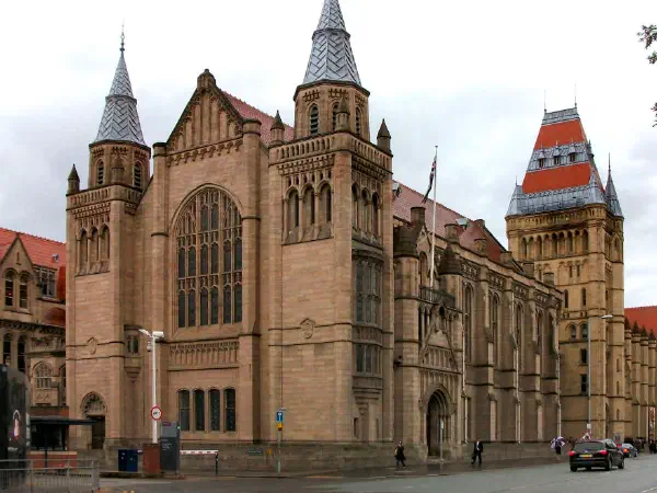 The red-roofed Victorian-style building with prominent spires stands on a cloudy day with pedestrians and cars nearby. — University of Manchester, Manchester