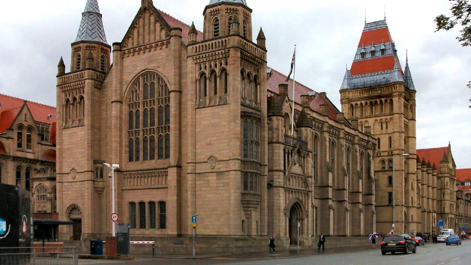 The red-roofed Victorian-style building with prominent spires stands on a cloudy day with pedestrians and cars nearby. — University of Manchester, Manchester