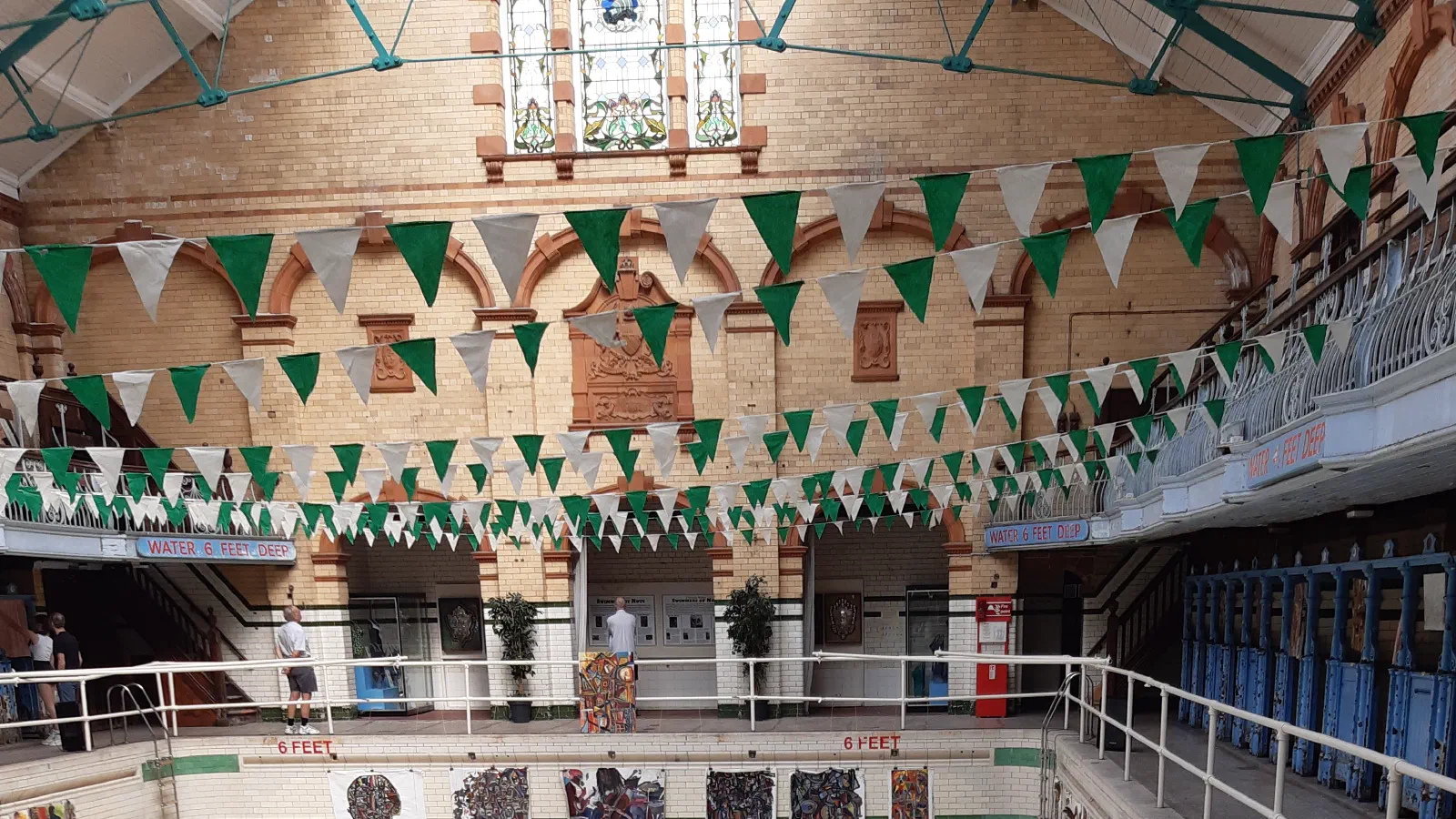 An empty swimming pool with green lane markers and colorful decorations, under a glass roof. — Victoria Baths, Manchester