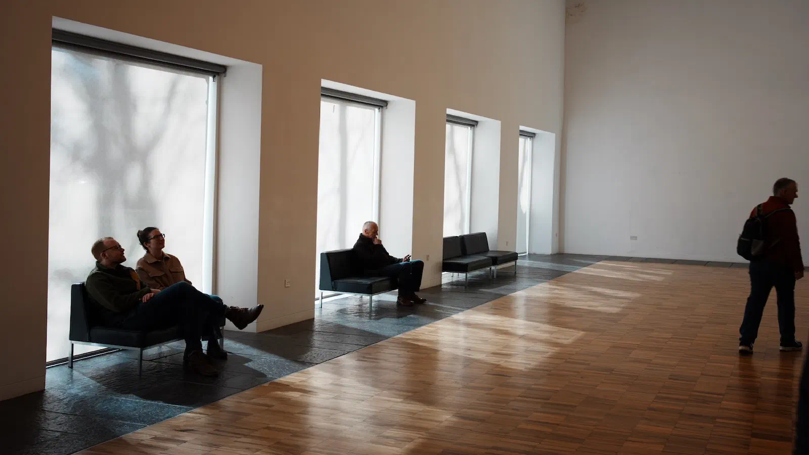 People sitting on benches near windows and another person walking in a spacious room with wooden floors. — Whitworth Art Gallery, Manchester