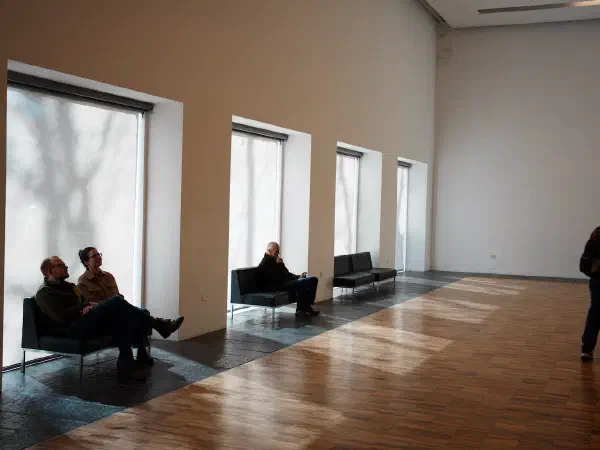 People sitting on benches near windows and another person walking in a spacious room with wooden floors. — Whitworth Art Gallery, Manchester