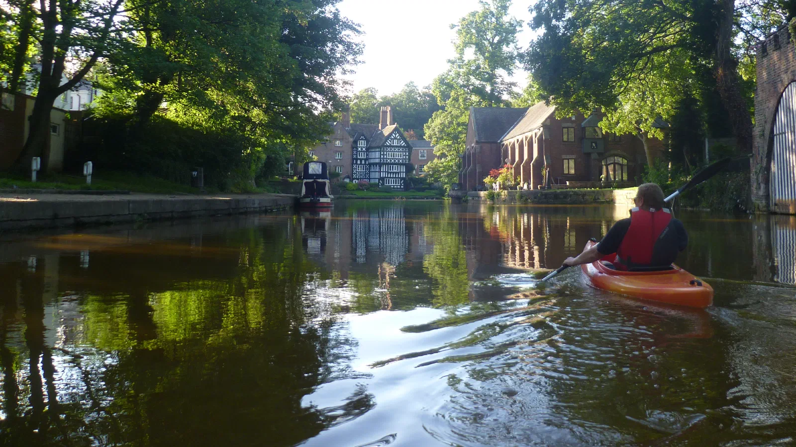 Person paddles kayak on a calm canal lined by greenery under bright light. — Worsley Village, Manchester