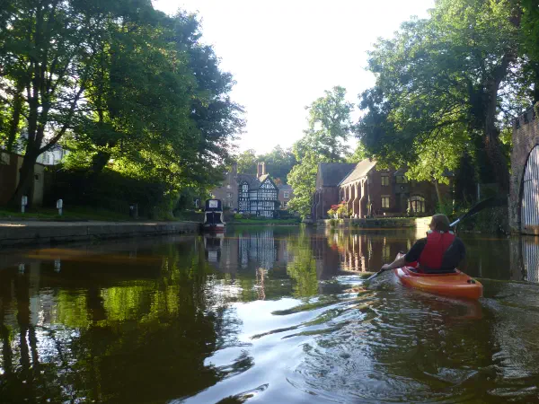 Person paddles kayak on a calm canal lined by greenery under bright light. — Worsley Village, Manchester